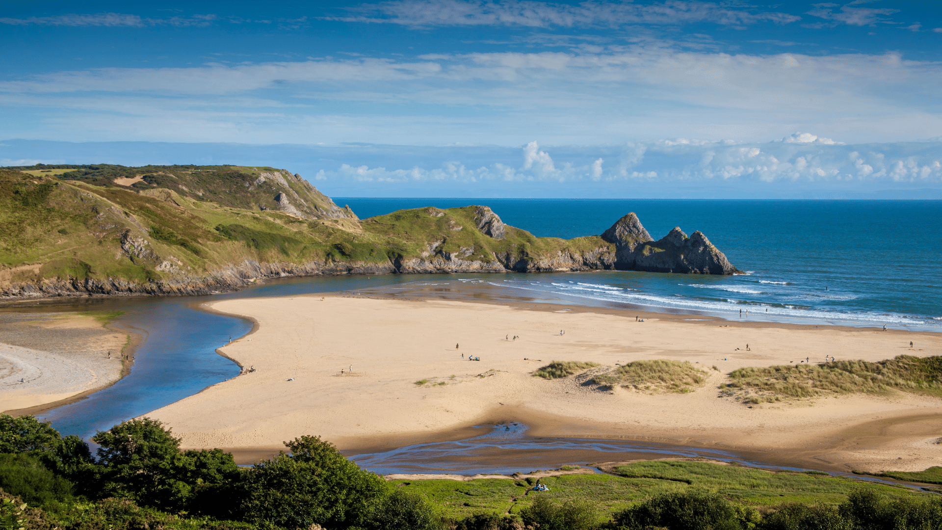 A sandy beach with grassy cliffs and blue sea under a partly cloudy sky.