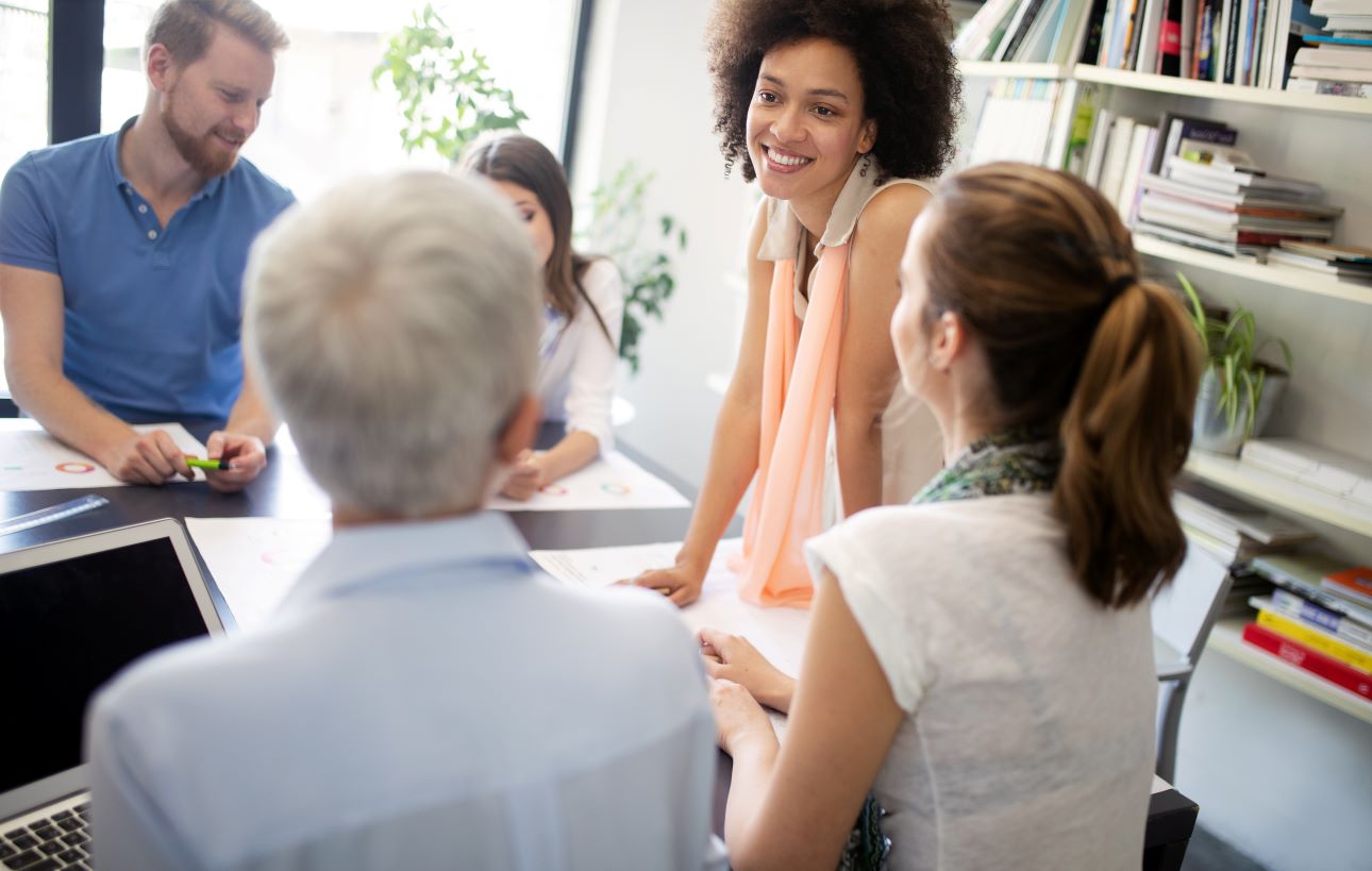 Four people sit and talk around a table in a casual office meeting.